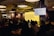 A speaker stands at a podium in a dimly lit room with an audience seated at tables. A large screen displays text about imposter syndrome. The room has industrial-style lighting and brick walls, creating a cozy atmosphere.