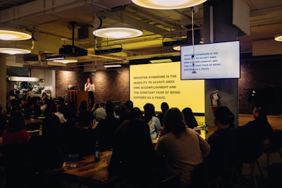 A speaker stands at a podium in a dimly lit room with an audience seated at tables. A large screen displays text about imposter syndrome. The room has industrial-style lighting and brick walls, creating a cozy atmosphere.