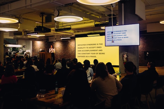 A speaker stands at a podium in a dimly lit room with an audience seated at tables. A large screen displays text about imposter syndrome. The room has industrial-style lighting and brick walls, creating a cozy atmosphere.