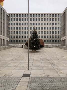 A large, modern office building with a grid-like pattern of windows stands behind an open courtyard. In the center of the courtyard, there is a decorated Christmas tree with red and gold ornaments. A tall flagpole is positioned in the foreground of the image.