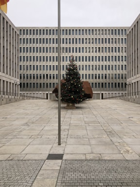 A large, modern office building with a grid-like pattern of windows stands behind an open courtyard. In the center of the courtyard, there is a decorated Christmas tree with red and gold ornaments. A tall flagpole is positioned in the foreground of the image.