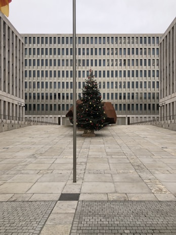 A large, modern office building with a grid-like pattern of windows stands behind an open courtyard. In the center of the courtyard, there is a decorated Christmas tree with red and gold ornaments. A tall flagpole is positioned in the foreground of the image.