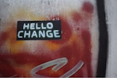 A vibrant street art mural featuring the words 'HELLO CHANGE' in bold white letters on a dark rectangular background. The backdrop is a dynamic blend of red and orange hues with patches of black and a hint of yellow, creating a sense of movement and transition.