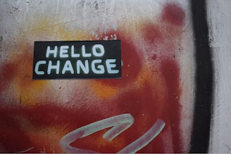 A vibrant street art mural featuring the words 'HELLO CHANGE' in bold white letters on a dark rectangular background. The backdrop is a dynamic blend of red and orange hues with patches of black and a hint of yellow, creating a sense of movement and transition.