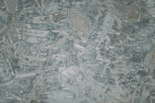 An abstract pattern with interconnected lines and shapes reminiscent of a topographical map. The texture is uneven with a mix of light and dark patches adding depth.