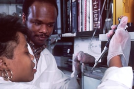 Two scientists are working together in a laboratory setting. One is holding a pipette and the other is closely observing. Both are wearing white lab coats and gloves, indicating they are conducting an experiment or research task. Shelves filled with books and equipment are visible in the background.