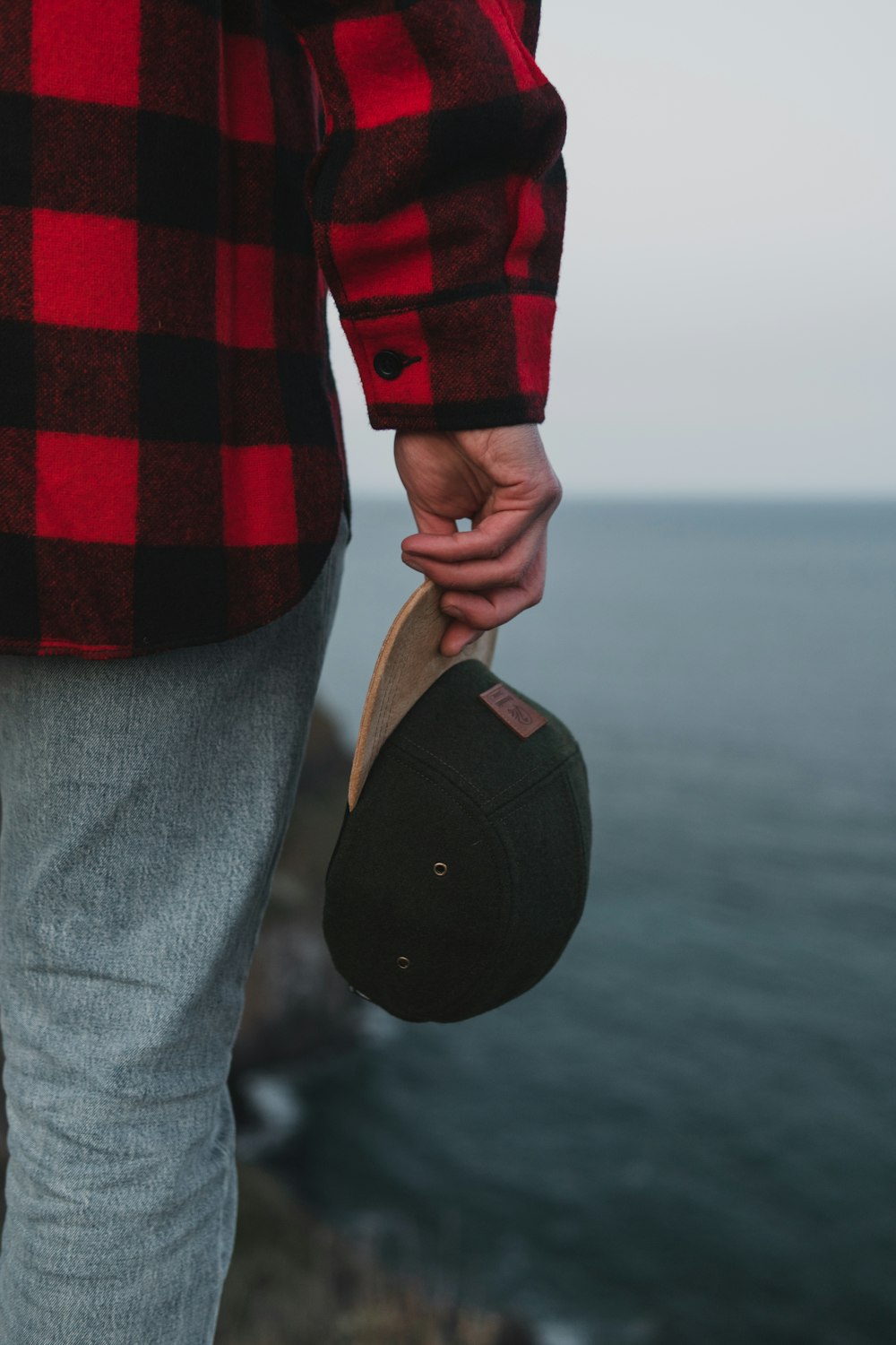 person wearing red and black long-sleeved shirt holding black and brown fitted cap while standing on cliff viewing body of water