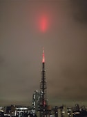 A tall broadcasting tower is illuminated with red lights, reaching up into a cloudy, overcast sky. The tower is situated in an urban environment, surrounded by a variety of buildings, including skyscrapers and smaller structures.