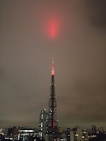A tall broadcasting tower is illuminated with red lights, reaching up into a cloudy, overcast sky. The tower is situated in an urban environment, surrounded by a variety of buildings, including skyscrapers and smaller structures.