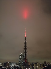 A tall broadcasting tower is illuminated with red lights, reaching up into a cloudy, overcast sky. The tower is situated in an urban environment, surrounded by a variety of buildings, including skyscrapers and smaller structures.