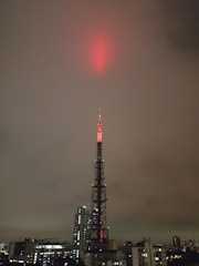 A tall broadcasting tower is illuminated with red lights, reaching up into a cloudy, overcast sky. The tower is situated in an urban environment, surrounded by a variety of buildings, including skyscrapers and smaller structures.