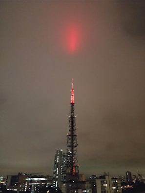 A tall broadcasting tower is illuminated with red lights, reaching up into a cloudy, overcast sky. The tower is situated in an urban environment, surrounded by a variety of buildings, including skyscrapers and smaller structures.