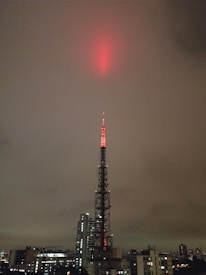 A tall broadcasting tower is illuminated with red lights, reaching up into a cloudy, overcast sky. The tower is situated in an urban environment, surrounded by a variety of buildings, including skyscrapers and smaller structures.