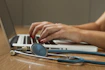 Hands typing on a laptop with a stethoscope resting on the wooden table beside it. The setting suggests a work environment that combines technology with healthcare.
