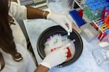A person wearing a lab coat and gloves is handling a container filled with test tubes and ice. The surrounding area shows laboratory equipment, including racks filled with colorful test tubes and other scientific materials.