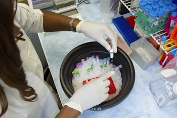 A person wearing a lab coat and gloves is handling a container filled with test tubes and ice. The surrounding area shows laboratory equipment, including racks filled with colorful test tubes and other scientific materials.