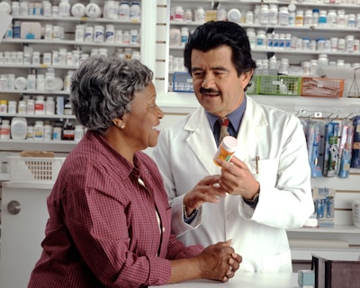 A pharmacist in a white coat talks to a customer in a pharmacy. Shelves filled with various medications are visible in the background. The pharmacist is holding a prescription bottle and appears to be explaining something to the customer, who is smiling and attentively listening.