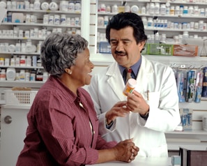 A pharmacist in a white coat talks to a customer in a pharmacy. Shelves filled with various medications are visible in the background. The pharmacist is holding a prescription bottle and appears to be explaining something to the customer, who is smiling and attentively listening.
