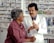 A pharmacist in a white coat talks to a customer in a pharmacy. Shelves filled with various medications are visible in the background. The pharmacist is holding a prescription bottle and appears to be explaining something to the customer, who is smiling and attentively listening.