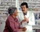 A pharmacist in a white coat talks to a customer in a pharmacy. Shelves filled with various medications are visible in the background. The pharmacist is holding a prescription bottle and appears to be explaining something to the customer, who is smiling and attentively listening.