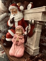 A festive scene featuring a Santa Claus figurine in a classic red suit with a white beard, holding a finger to his lips in a gesture of secrecy. Next to him is a child figurine in a pink gown, kneeling and praying. The setting includes a decorative fireplace with stockings and a small antique clock on the mantle, surrounded by Christmas-themed decorations.
