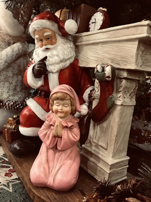 A festive scene featuring a Santa Claus figurine in a classic red suit with a white beard, holding a finger to his lips in a gesture of secrecy. Next to him is a child figurine in a pink gown, kneeling and praying. The setting includes a decorative fireplace with stockings and a small antique clock on the mantle, surrounded by Christmas-themed decorations.