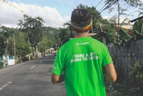A person wearing a bright green shirt with a motivational text stands on a road surrounded by lush greenery. The shirt reads 'Change a life, change the nation.' A colorful headband adorns their head. The road is empty, flanked by trees and plants, conveying a serene and peaceful setting.