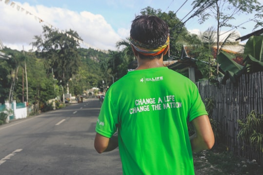 A person wearing a bright green shirt with a motivational text stands on a road surrounded by lush greenery. The shirt reads 'Change a life, change the nation.' A colorful headband adorns their head. The road is empty, flanked by trees and plants, conveying a serene and peaceful setting.