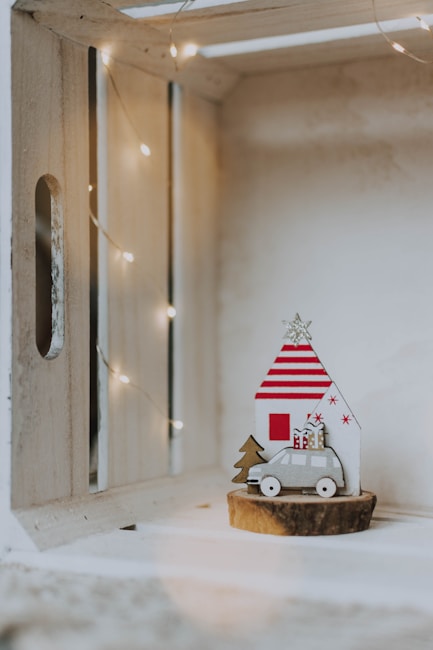 A small wooden shelf holds a festive holiday decoration. A miniature wooden car with gift boxes on top is parked in front of a stylized house with red stripes, a red door, and a silver star on the roof. A small wooden tree stands beside the car. Soft, warm string lights border the shelf, creating a cozy ambiance.