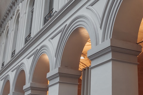 A series of elegant, classical arches are seen, each supported by robust columns. The architecture is ornate and detailed, featuring decorative moldings and clean lines that suggest a historical or formal building. Soft lighting illuminates the arches, creating gentle shadows and highlighting the structure's symmetry.