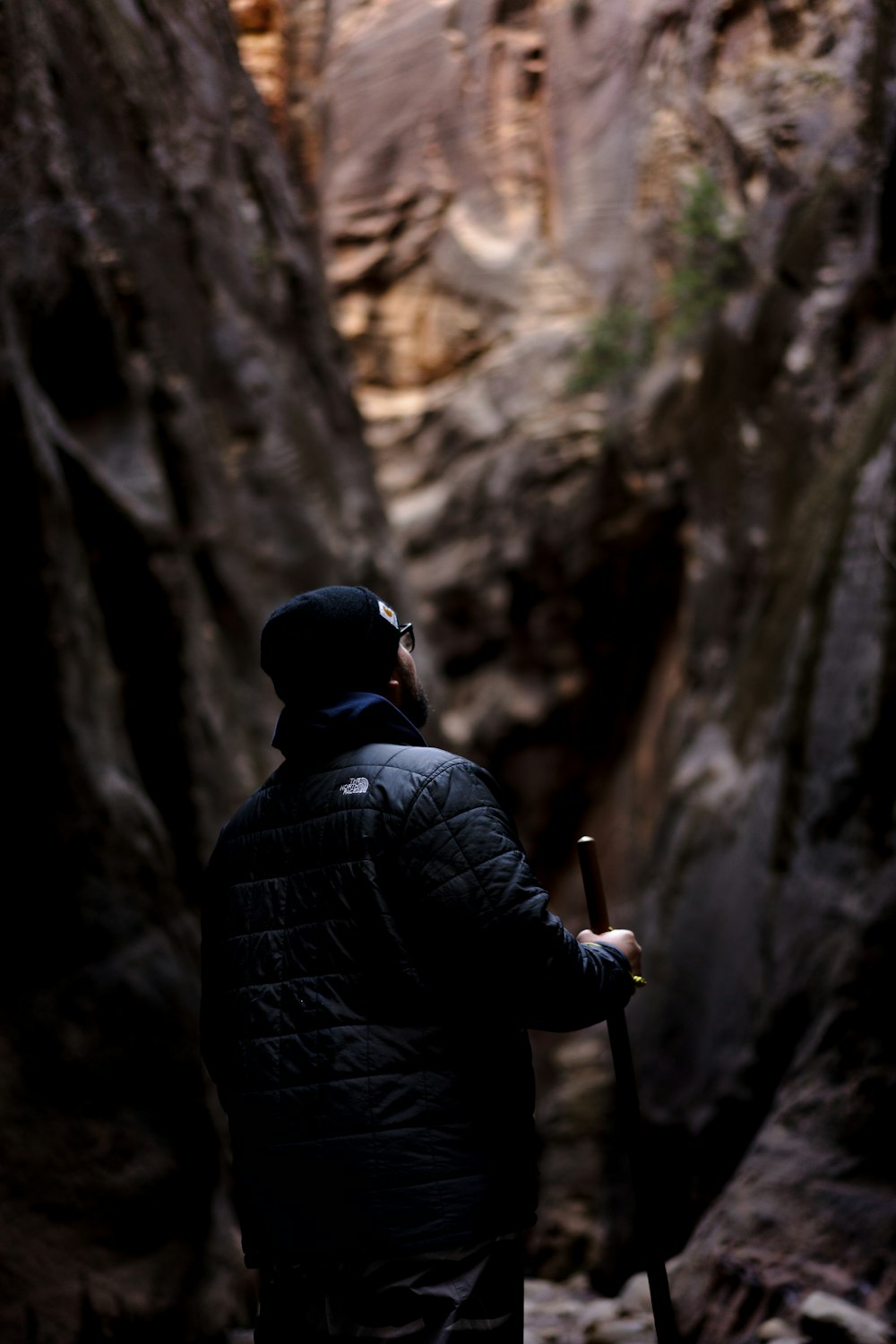 man wearing black jacket holding stick while standing near cave during daytime