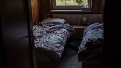 A dimly lit room with two single beds covered in rumpled bedding. There is a small wooden nightstand between the beds, which holds a few personal items like a candle and a tube of lotion. A window above the radiator provides a view of greenery outside, suggesting a peaceful, secluded setting.
