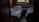 A dimly lit room with two single beds covered in rumpled bedding. There is a small wooden nightstand between the beds, which holds a few personal items like a candle and a tube of lotion. A window above the radiator provides a view of greenery outside, suggesting a peaceful, secluded setting.