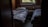 A dimly lit room with two single beds covered in rumpled bedding. There is a small wooden nightstand between the beds, which holds a few personal items like a candle and a tube of lotion. A window above the radiator provides a view of greenery outside, suggesting a peaceful, secluded setting.
