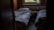 A dimly lit room with two single beds covered in rumpled bedding. There is a small wooden nightstand between the beds, which holds a few personal items like a candle and a tube of lotion. A window above the radiator provides a view of greenery outside, suggesting a peaceful, secluded setting.