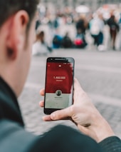 A person is holding a smartphone displaying a social media app with subscriber count. The screen shows 1,460,000 subscribers and features a red and white interface. The background is blurred with a crowd of people, suggesting an outdoor or public setting.