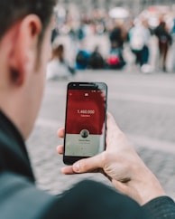 A person is holding a smartphone displaying a social media app with subscriber count. The screen shows 1,460,000 subscribers and features a red and white interface. The background is blurred with a crowd of people, suggesting an outdoor or public setting.