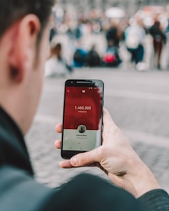 A person is holding a smartphone displaying a social media app with subscriber count. The screen shows 1,460,000 subscribers and features a red and white interface. The background is blurred with a crowd of people, suggesting an outdoor or public setting.