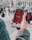 A hand holds a smartphone displaying a social media app with 118,000 subscribers on the screen. The image is set outdoors in a public plaza with multiple people blurred in the background.