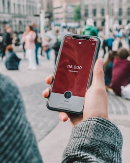 A hand holds a smartphone displaying a social media app with 118,000 subscribers on the screen. The image is set outdoors in a public plaza with multiple people blurred in the background.