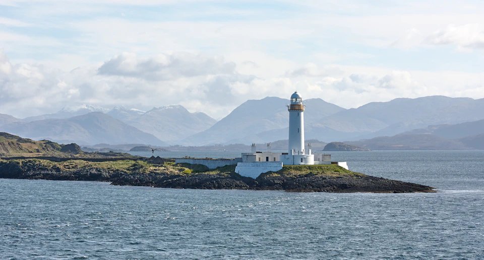 The image depicts a serene scene at a lighthouse on an island. The lighthouse, painted white with a yellow roof and standing tall against the sky, is surrounded by a rocky shoreline. The water surrounding the island is a deep blue color, reflecting the clear sky above. In the distance, mountains can be seen rising up from the horizon, adding depth to the scene.
