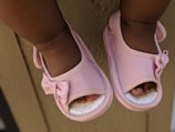 A close-up view of a child's feet wearing pink sandals adorned with small bows. The sandals have a soft pastel tone and fit snugly on the child's feet. The background has a neutral wooden or wall surface.