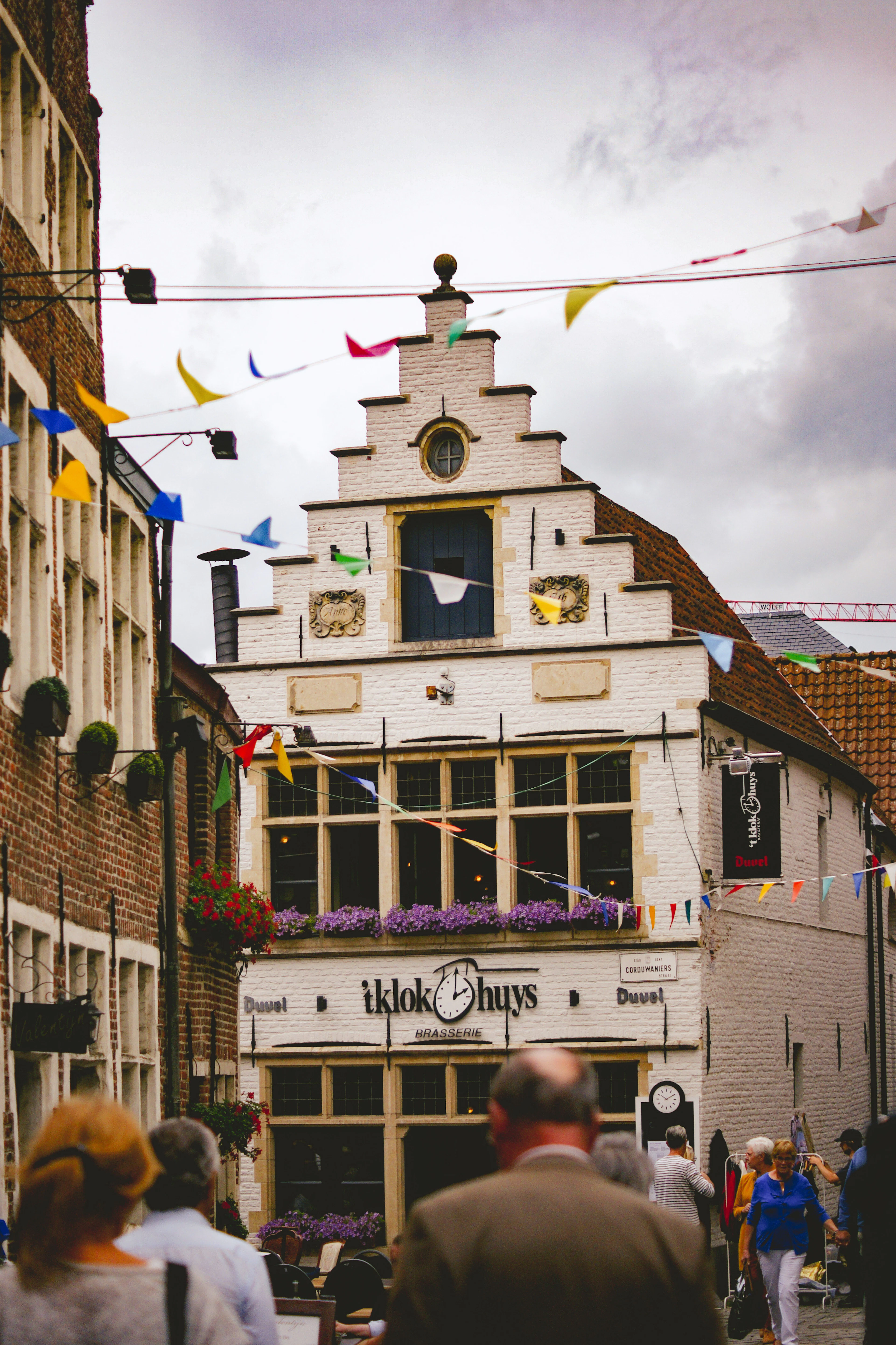 The white stepped-gable facade of the 't Klokhuys brasserie in Ghent's Patershol quarter, strung with coloured bunting and hung with purple window boxes, people at the tables below.