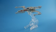 A model of a futuristic spacecraft is displayed mounted on a clear plastic stand against a plain blue background. The design is sleek with recognizable wings and engines extending from a central body.