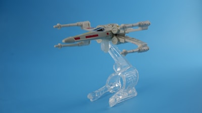 A model of a futuristic spacecraft is displayed mounted on a clear plastic stand against a plain blue background. The design is sleek with recognizable wings and engines extending from a central body.