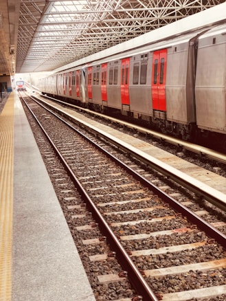 A modern train station platform with metal tracks and a stationary train. The train has a metallic body with red doors and windows. The platform is covered by a semi-transparent roof structure with metal beams, allowing natural light to filter through. The platform area is clean and well-maintained, with yellow tactile paving on the edge for safety.