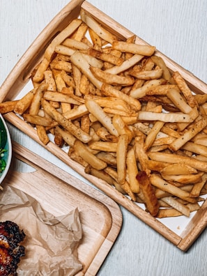 A wooden tray filled with a large amount of seasoned French fries. Next to the tray is a plate that has partially visible grilled meat with a dark, caramelized edge, served on a piece of parchment paper.