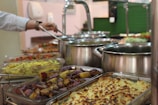 A buffet setup with a variety of dishes in trays and pots. The assortment includes baked potatoes, a yellow rice dish, and other vegetable preparations. A person is serving food with a ladle, and there are multiple stainless steel pots in the background.