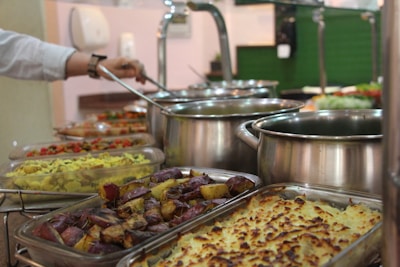 A buffet setup with a variety of dishes in trays and pots. The assortment includes baked potatoes, a yellow rice dish, and other vegetable preparations. A person is serving food with a ladle, and there are multiple stainless steel pots in the background.