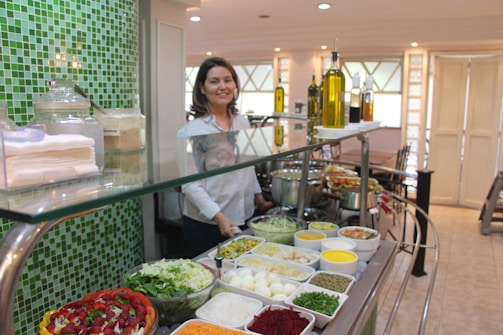 A person stands behind a glass counter featuring an array of colorful salads and condiments in bowls and trays. The counter is topped with bottles of oil and utensils. The background shows a well-lit room with light-colored walls and glass windows, giving a clean and inviting atmosphere.